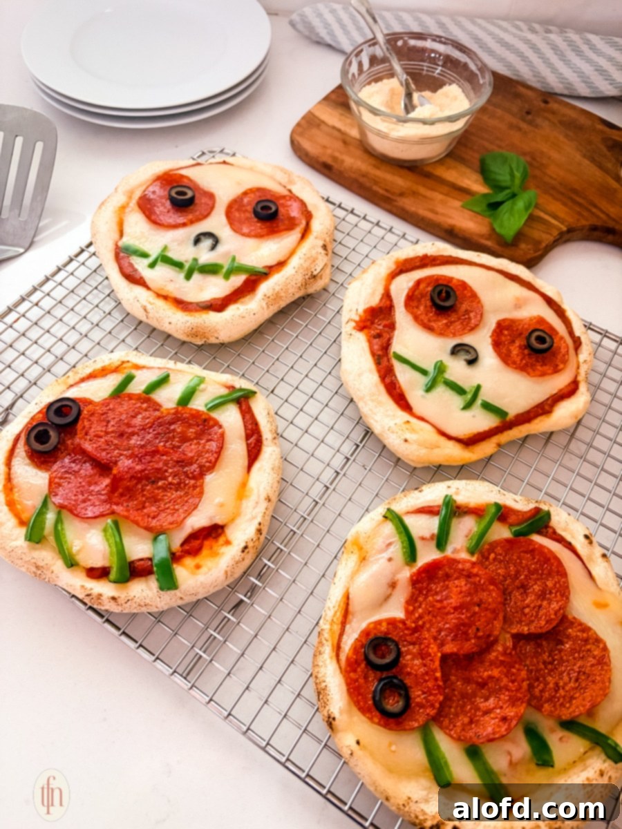 Monster Mash Pizza 3 Four baked personal Halloween pizzas are shown on a wire cooling rack. Two are decorated as skulls with sliced olives for eyes and green peppers for a mouth, and two are decorated as spiders with pepperoni for the body and green pepper slices for legs.