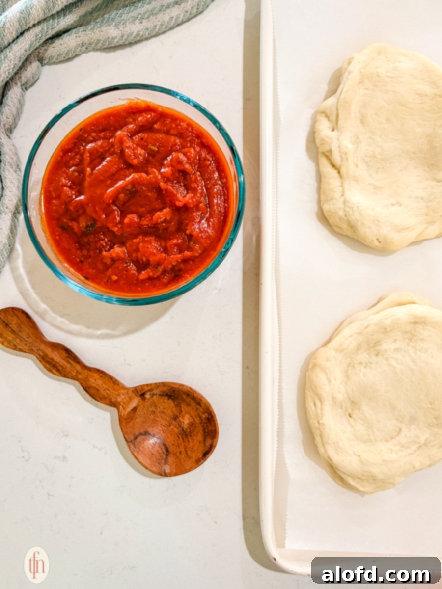 Monster Mash Pizza 7 A bowl of marinara sauce and two rounds of pizza dough on a baking sheet lined with parchment paper are shown from above. A wooden spoon rests near the sauce bowl.