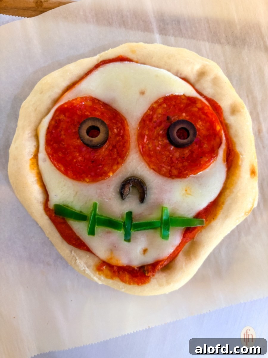 Monster Mash Pizza 8 A close-up shot of an unbaked Halloween-themed personal pizza shaped like a skull. It has a marinara sauce base, mozzarella cheese, pepperoni for the eyes, sliced black olives for the pupils and nose, and small strips of green bell pepper for the mouth stitches.