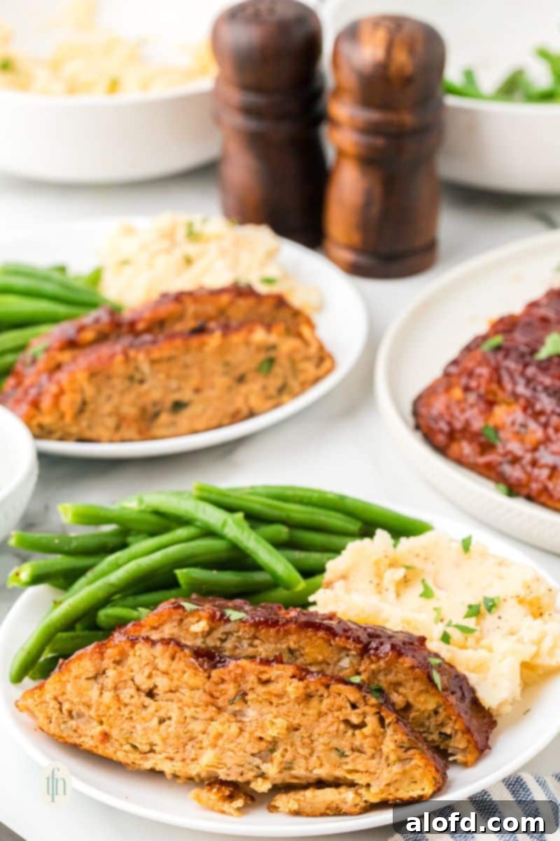 Savory Chicken Meatloaf 2 An eye-level shot showing two plates of glazed chicken meatloaf, mashed potatoes, and green beans, ready to be served. Two wooden spice shakers are visible in the background.