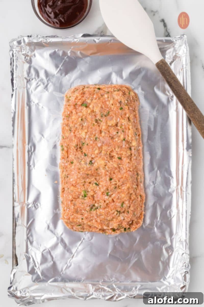 Savory Chicken Meatloaf 13 An overhead shot of the raw chicken meatloaf mixture shaped into a loaf on a foil-lined baking sheet. A white spatula is resting next to it, and a small bowl of glaze is visible at the top.