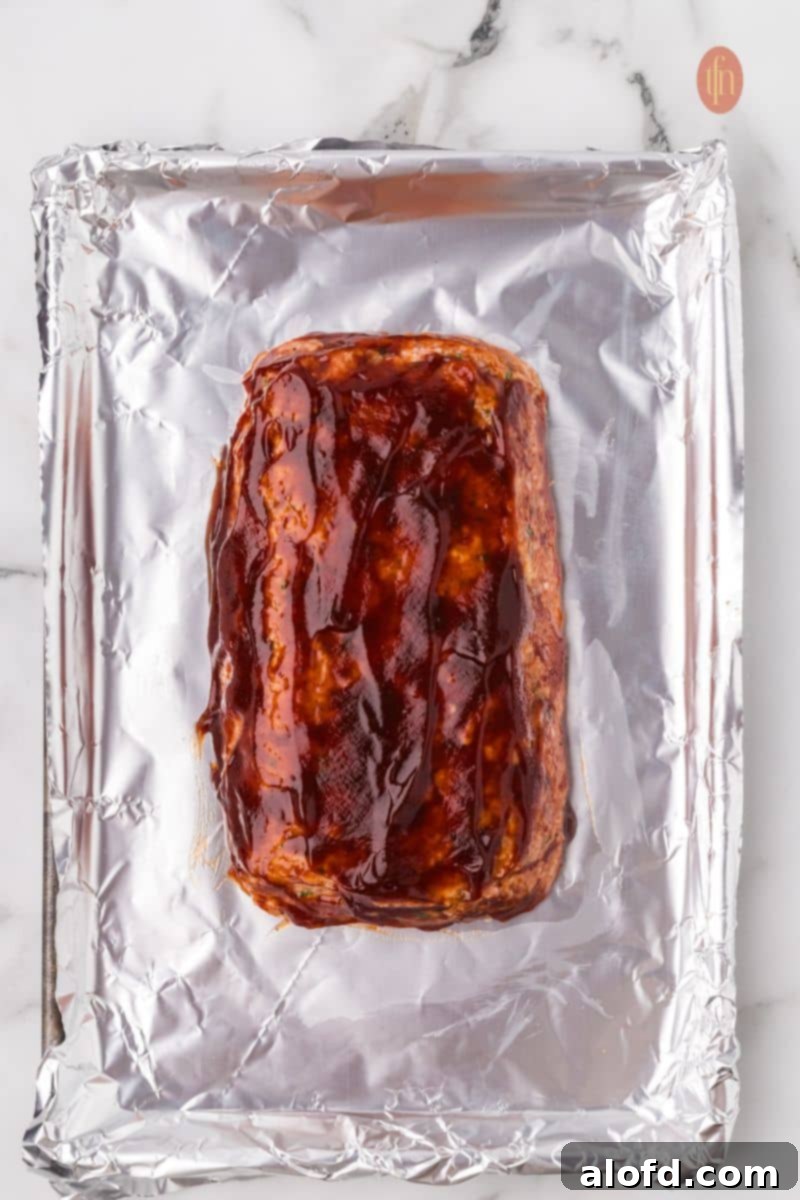 Savory Chicken Meatloaf 14 An overhead shot of the raw chicken meatloaf on a foil-lined baking sheet, now fully brushed with a generous layer of thick glaze before baking.