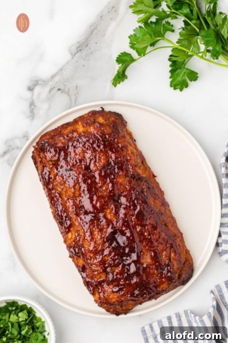 Savory Chicken Meatloaf 16 An overhead shot of a whole chicken meatloaf on a white platter. It is covered in a glossy, dark red glaze. A sprig of parsley and a blue and white striped towel are nearby.