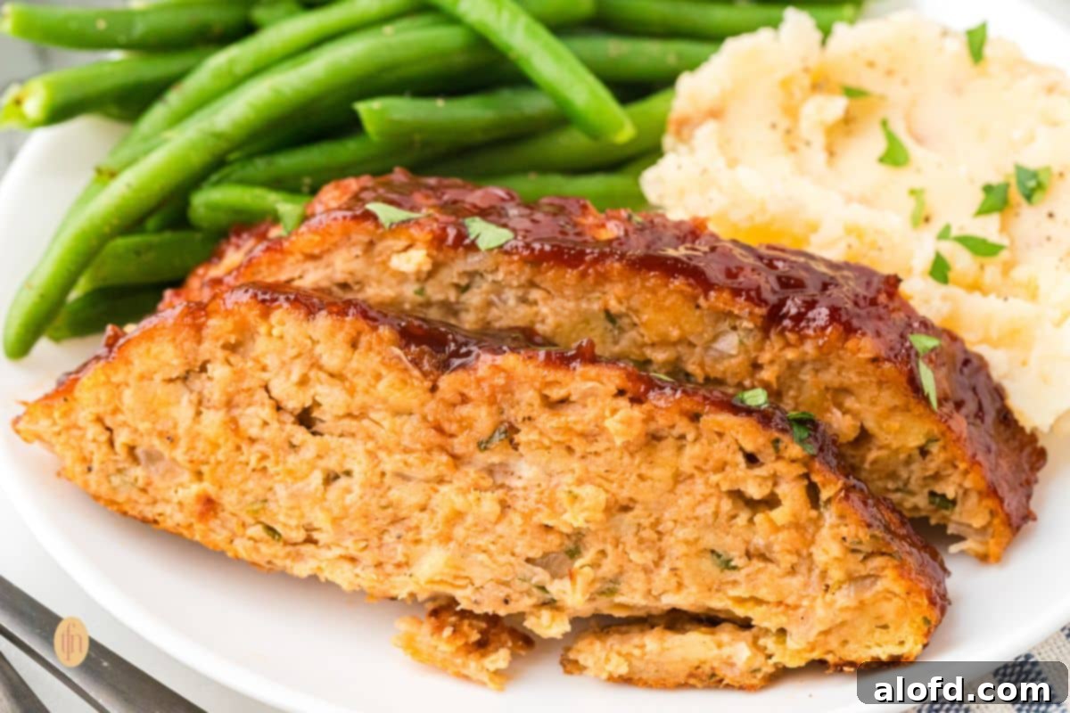 Savory Chicken Meatloaf 17 A wide, close-up shot of two slices of glazed chicken meatloaf served on a white plate with green beans and mashed potatoes.