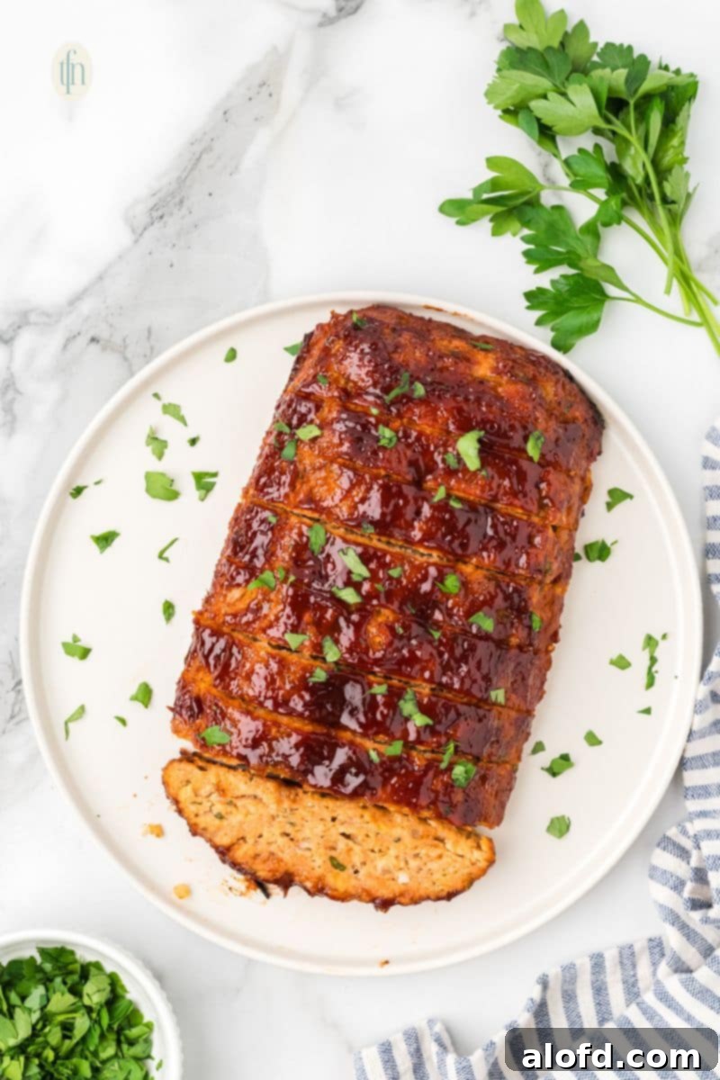 Savory Chicken Meatloaf 3 An overhead shot of a glazed chicken meatloaf on a white platter, already sliced into about eight thick pieces and garnished with chopped parsley.