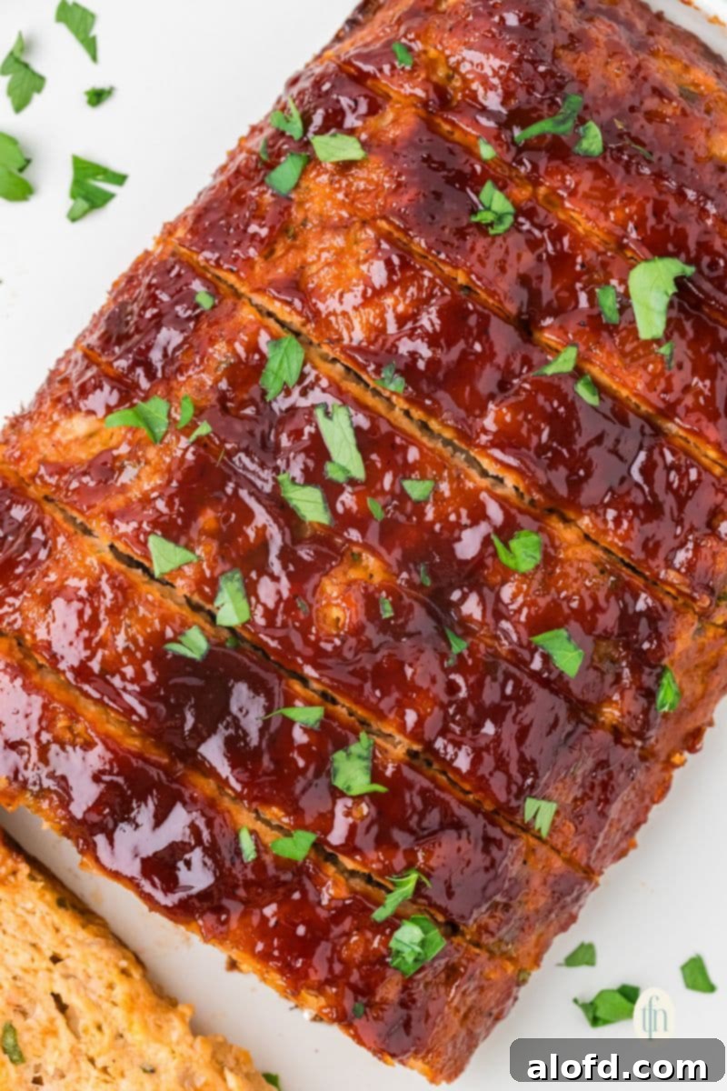 Savory Chicken Meatloaf 4 A close-up, overhead view of a sliced chicken meatloaf. The top is covered in a thick, shiny red glaze and sprinkled with chopped parsley.