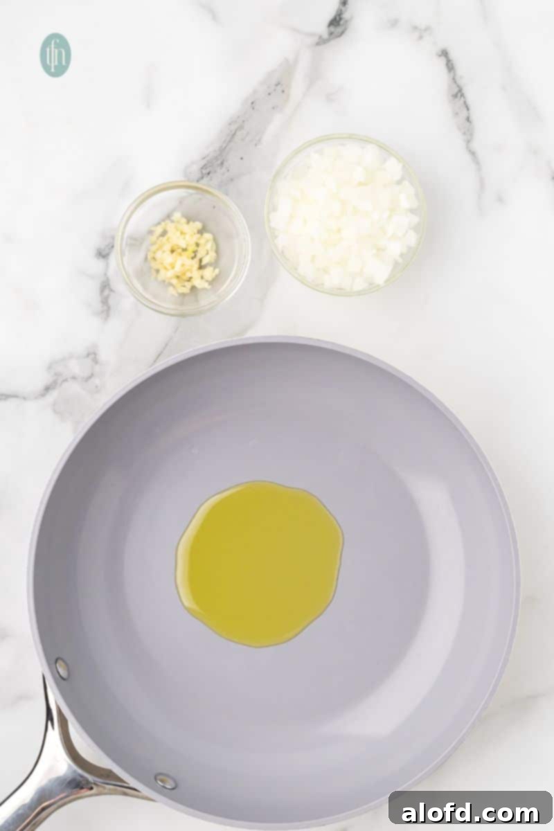 Savory Chicken Meatloaf 6 An overhead shot of a gray nonstick skillet with a pool of olive oil. Small glass bowls of chopped garlic and diced onion are visible on the marble counter.
