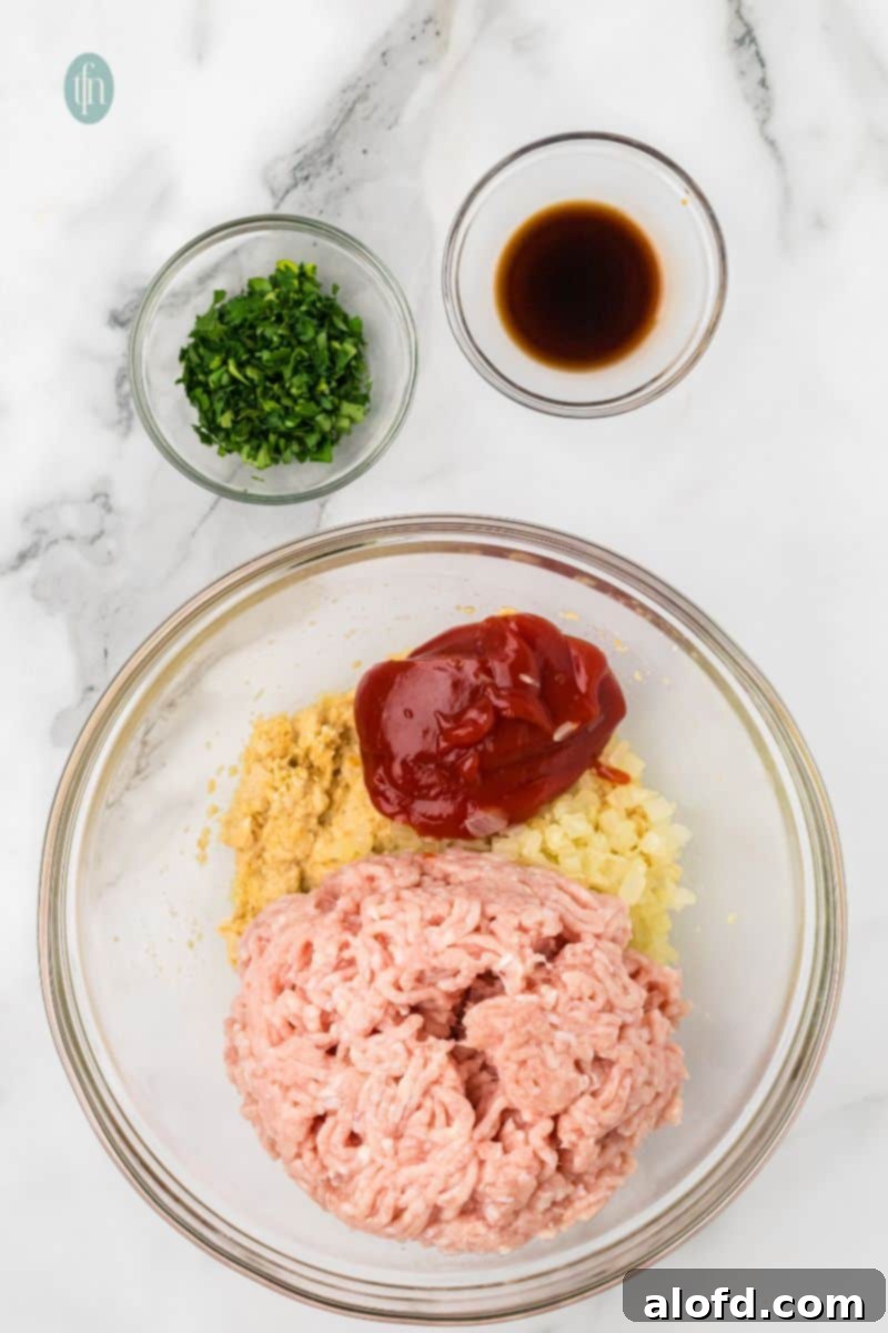 Savory Chicken Meatloaf 10 A large glass bowl is filled with raw ground chicken, the soaked breadcrumb mixture, sautéed onions/garlic, a dollop of ketchup, and a small amount of dark liquid (likely Worcestershire or soy sauce). A bowl of chopped parsley is on the side.