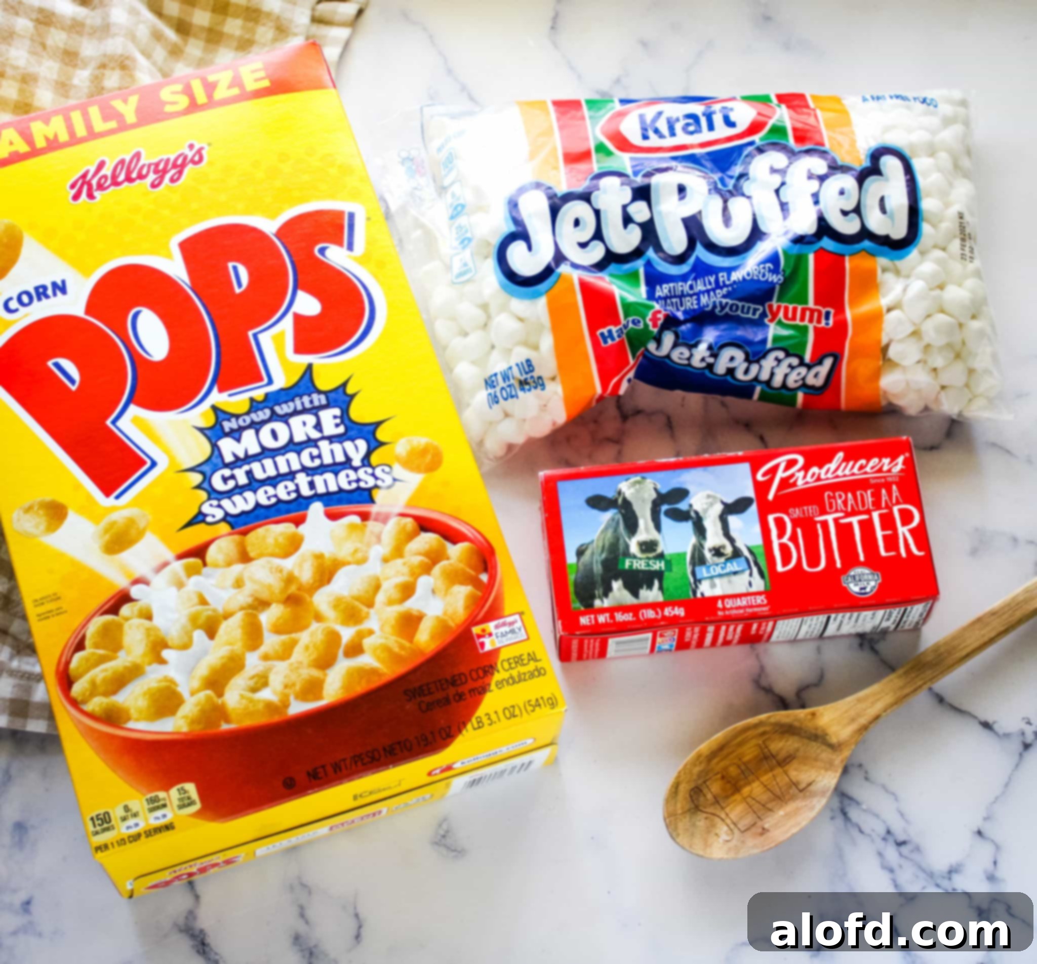 Ingredients for Corn Pops treats: a bowl of golden Corn Pops cereal, a bag of fluffy marshmallows, and a stick of butter, neatly arranged on a wooden surface.