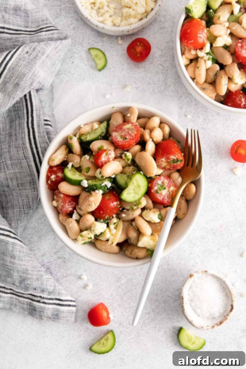 Tuscan white bean salad with a fork in a white bowl with cloth, another bowl of tuscan salad, and chopped tomatoes ad cucumbers at the background.