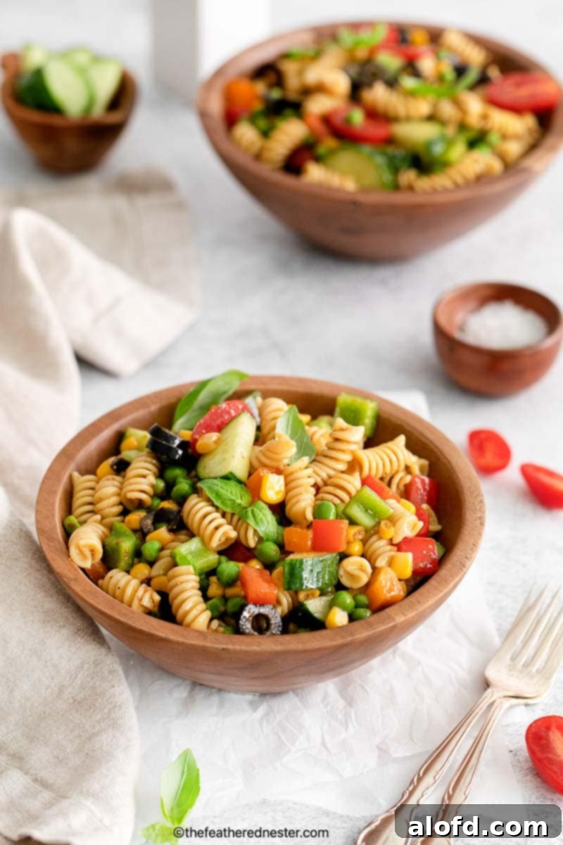 zesty Italian pasta salad and silverwares on a wood bowl with another bowl of salad and salt, cucumber, and cherry tomatoes placed on a white cloth.