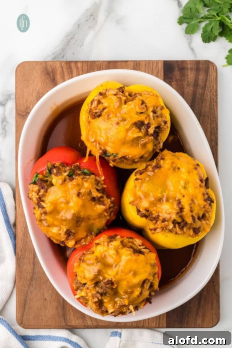 Loaded Taco Peppers 16 An overhead shot of four baked stuffed bell peppers in a white oval baking dish, sitting on a wooden cutting board with a blue and white striped towel. The peppers are topped with melted cheese and sit in a pool of rich, red sauce, creating a rustic and inviting scene.