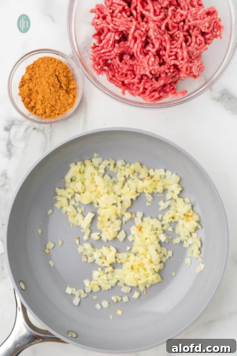 Loaded Taco Peppers 7 Diced onions and garlic are sautéing in a gray pan. Ingredients for the filling, including a bowl of ground beef and a bowl of taco seasoning, are visible on the counter above the pan, indicating the next steps in preparation.