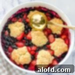 a white casserole dish with strawberry and blueberry cobbler and a gold serving spoon.