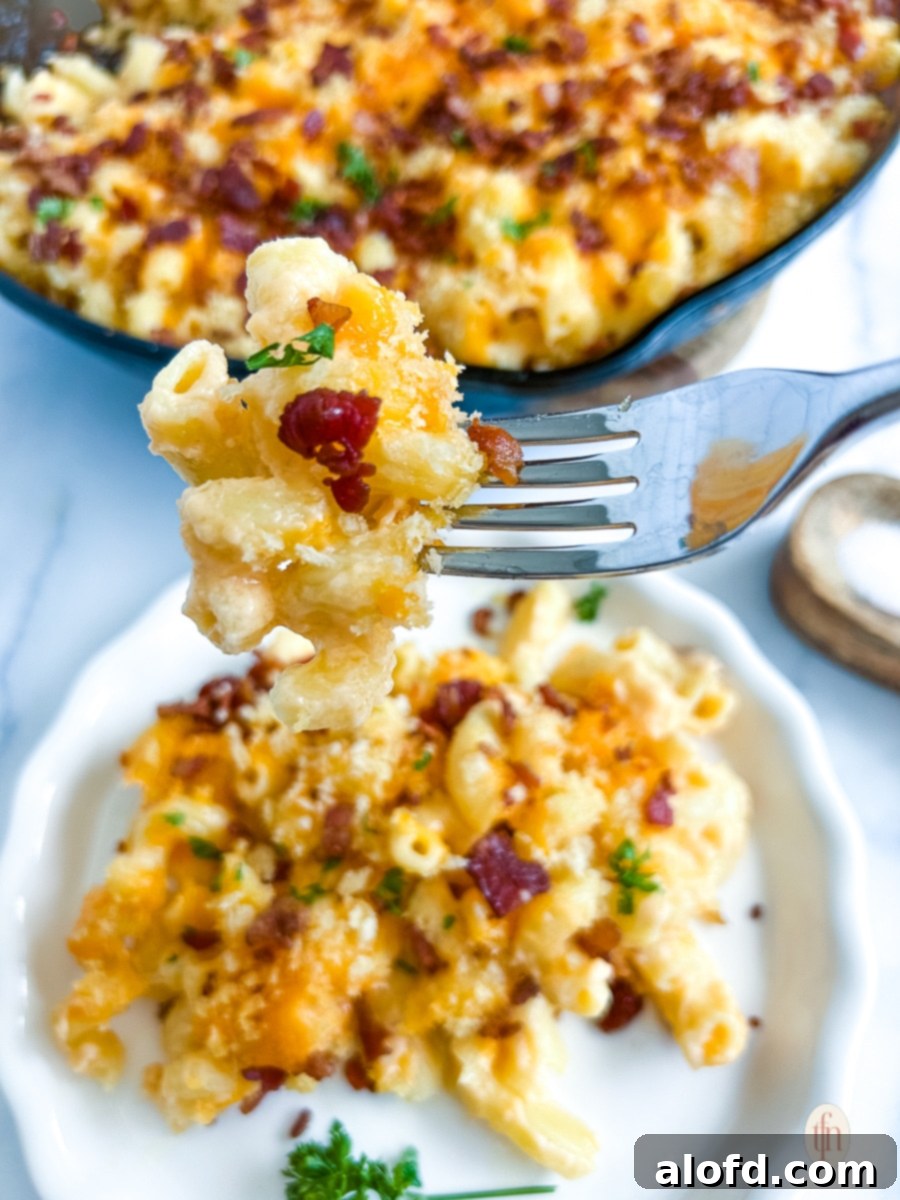 A forkful of smoked mac and cheese, shown close up above a plated serving. A large skillet of mac and cheese with bacon is visible in the background.