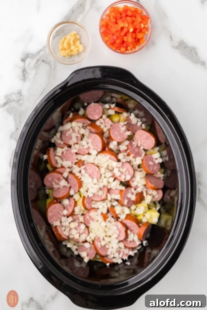Slow-Cooker Sausage and Potato Delight 6 Crockpot with potatoes, onions, and sausage, with bell peppers and garlic next to it.